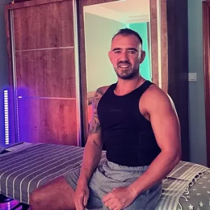 A man sitting on a massage table in a cozy home environment, smiling at the camera. The setup suggests a relaxing at-home massage session.